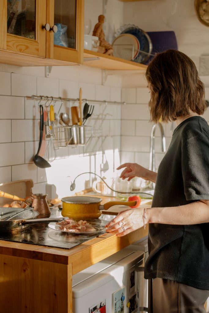 woman-in-gray-long-sleeve-shirt-standing-in-front-of-kitchen-sink-4781442 A woman cooks breakfast in a sunlit kitchen, featuring bacon and kitchenware.